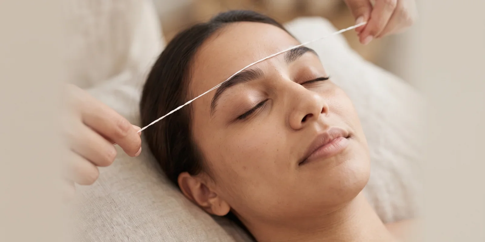A woman lying back with her eyes closed while a twisted cotton thread is held along the top edge of her eyebrow
